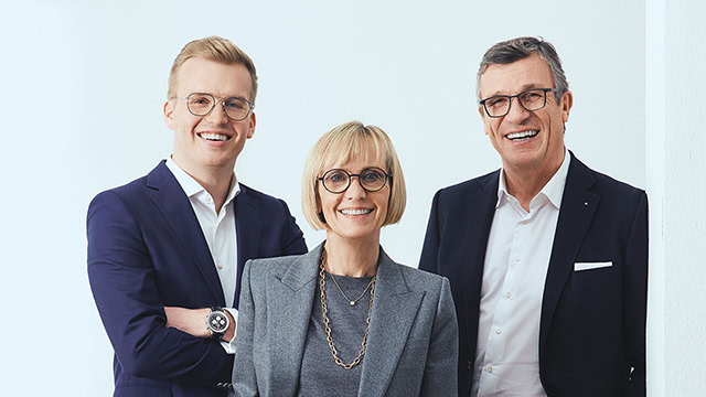 Birgit Matt, Michael Matt und Maximilian Matt stehen in Anzügen vor einer hellblauen Wand und lächeln freundlich in die Kamera.