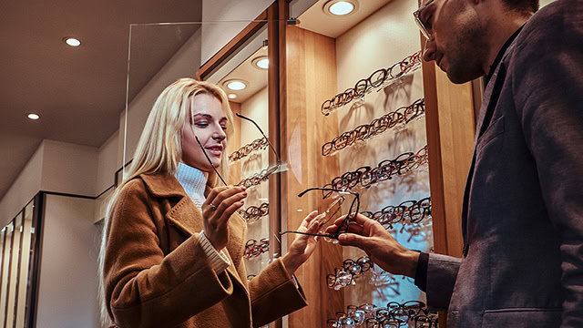 Eine freundliche Verkaufssituation bei einem Optiker: Ein Verkäufer und eine Kundin stehen vor einer geöffneten Glasvitrine, während die Kundin eine Brille gegen eine andere tauscht, die 									ihr der Verkäufer reicht.