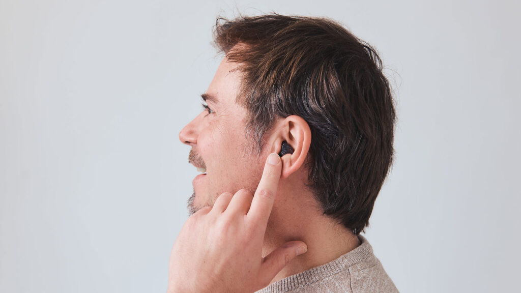 Mann mit Bart und braunen Haaren blickt zur Seite und deutet mit dem Zeigefinger auf ein Hörgerät in seinem linken Ohr